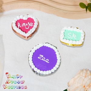 Peut inclure: Trois miroirs compacts décoratifs avec des motifs de gâteaux. L'un est en forme de cœur avec "Kaye" en blanc, un autre est rectangulaire avec "SR", et le troisième est rond avec "Zoe". Chacun a une bordure de glaçage blanc et un cadre en métal doré.