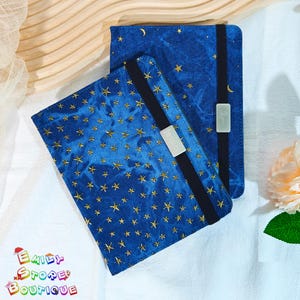 May include: Two blue fabric book covers with gold star patterns. Each cover has a black elastic band and a white rectangular clasp. One cover has the name "Jessica" printed on the clasp.