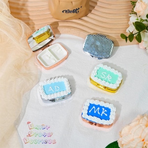 Può includere: Un assortimento di piccoli portapillole decorativi. Alcuni sono argentati o dorati con coperchi a fantasia o a specchio. Altri sono progettati per assomigliare a torte in miniatura, con glassa blu, verde e bianca e iniziali. È visibile una borsa marrone con la scritta "Best".