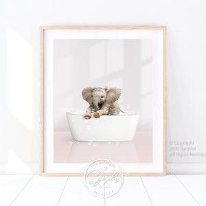May include: A cute baby elephant taking a bath in a white bathtub with bubbles. The elephant is sitting in the tub with its trunk up and its ears out. The background is a light pink color.