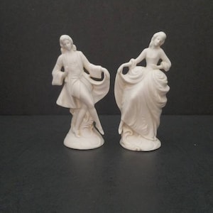 May include: A pair of white porcelain figurines depicting a man and a woman in a dancing pose. The man is wearing a long coat and the woman is wearing a long dress.