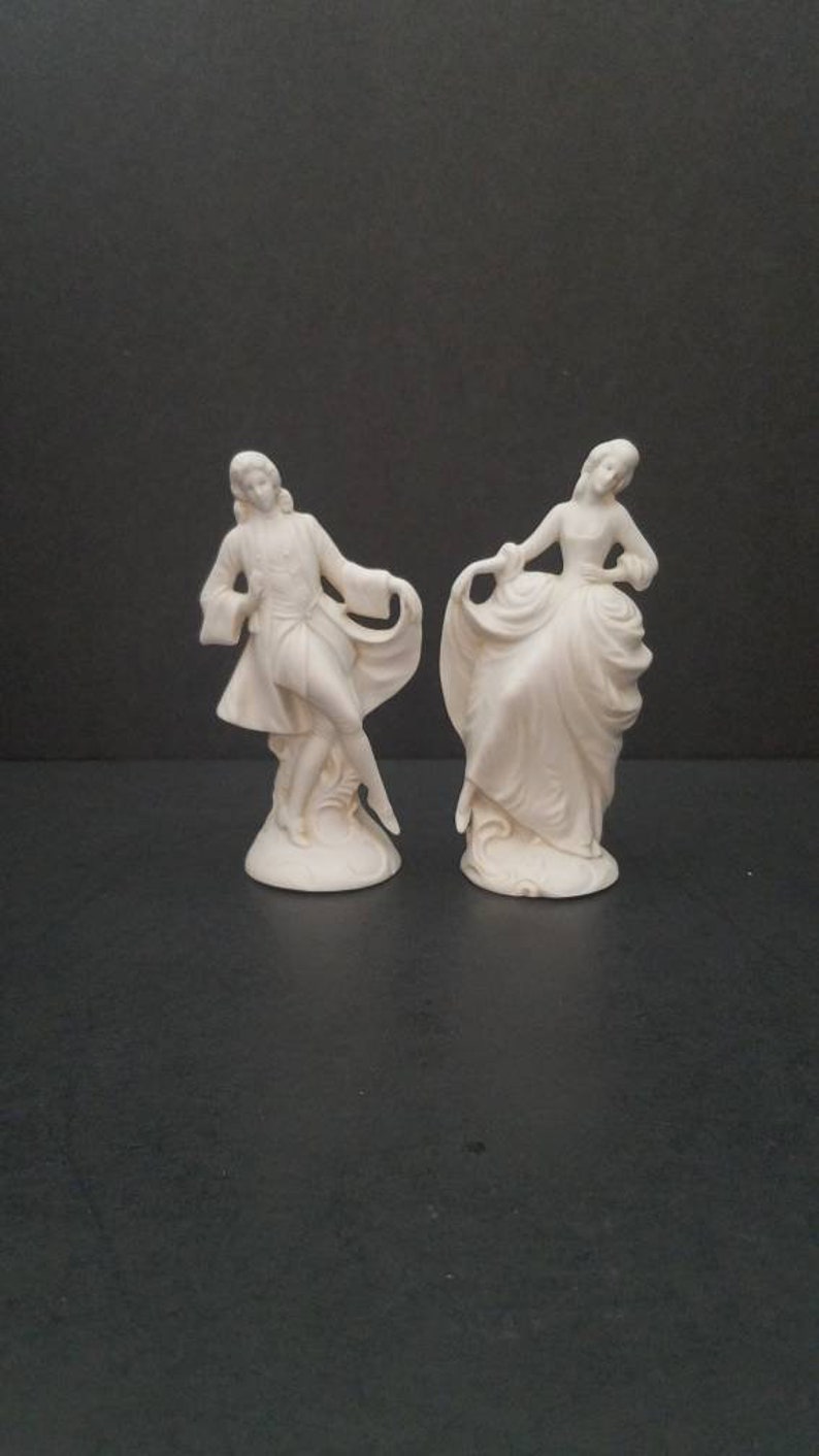 May include: A pair of white porcelain figurines depicting a man and a woman in a dancing pose. The man is wearing a long coat and the woman is wearing a long dress.