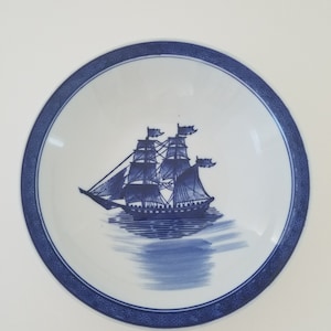 May include: A white ceramic bowl with a blue and white design of a ship sailing on the water. The bowl has a blue and white border with a geometric pattern.