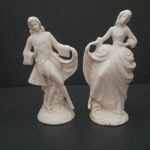 May include: A pair of white porcelain figurines depicting a man and woman in 18th-century attire. The man is wearing a long coat and the woman is wearing a dress with a large skirt. Both figurines are standing on a base.