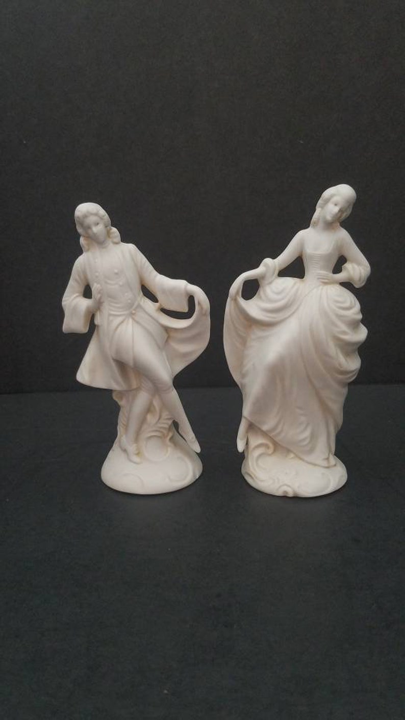 May include: A pair of white porcelain figurines depicting a man and woman in 18th-century attire. The man is wearing a long coat and the woman is wearing a dress with a large skirt. Both figurines are standing on a base.