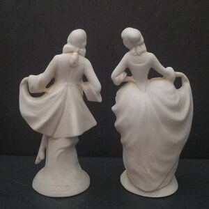 May include: A pair of white porcelain figurines depicting a man and a woman in 18th-century attire. The man is wearing a long coat and the woman is wearing a dress with a large skirt. Both figurines are standing on a base with the number "3229" printed on it.