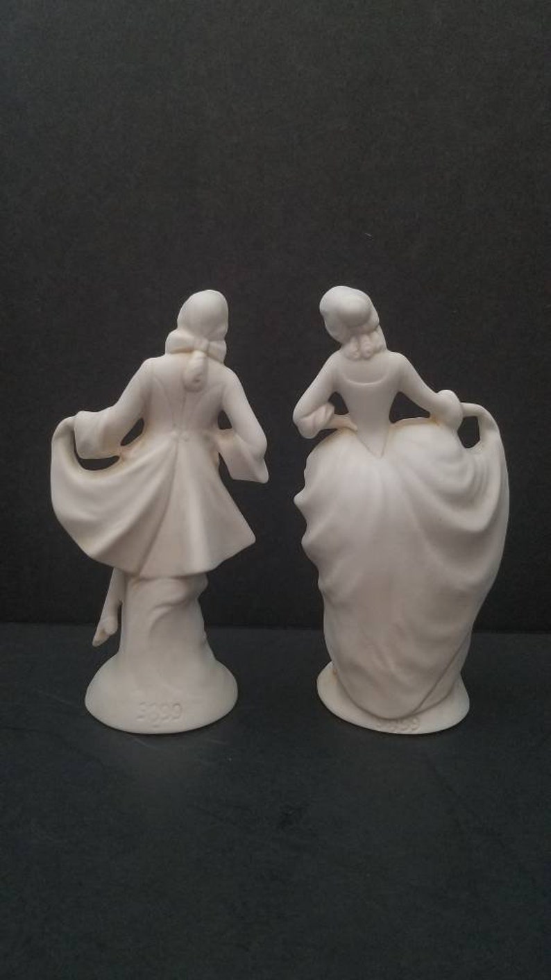 May include: A pair of white porcelain figurines depicting a man and a woman in 18th-century attire. The man is wearing a long coat and the woman is wearing a dress with a large skirt. Both figurines are standing on a base with the number "3229" printed on it.