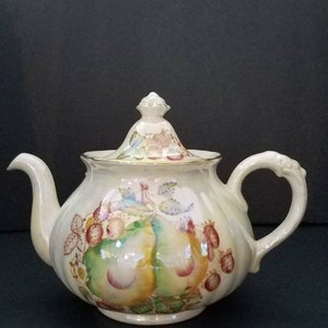 May include: A white ceramic teapot with a floral design featuring pears and strawberries. The teapot has a lid with a knob and a handle on the side.