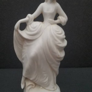 May include: A white porcelain figurine of a woman in a flowing gown. The figurine is standing on a base with a decorative design.