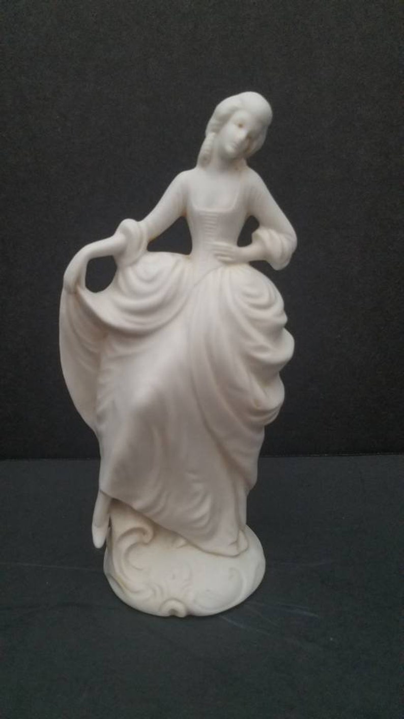 May include: A white porcelain figurine of a woman in a flowing gown. The figurine is standing on a base with a decorative design.