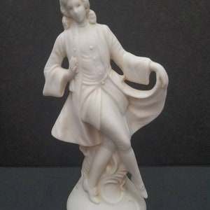 May include: A white porcelain figurine of a person in a long coat with a flowing cape. The figurine is standing on a round base.