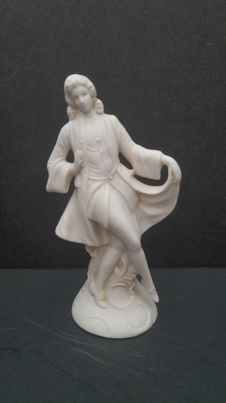 May include: A white porcelain figurine of a person in a long coat with a flowing cape. The figurine is standing on a round base.