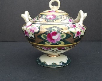 May include: A white porcelain sugar bowl with a lid and two handles. The bowl is decorated with pink and green floral designs and gold accents.