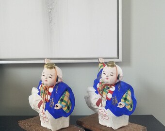 May include: Two porcelain figurines of children wearing traditional Japanese clothing. The figurines are painted in bright colors, including blue, yellow, red, and white. They are standing on wooden bases.