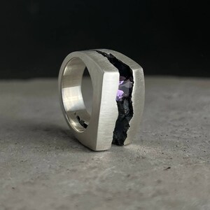 May include: A silver ring with a black crack down the center. A purple gemstone is set in the crack.