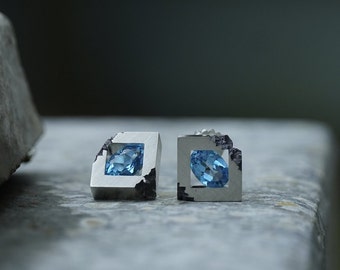 May include: A pair of silver earrings with a blue gemstone set in a square frame. The frame has a rough, textured edge.