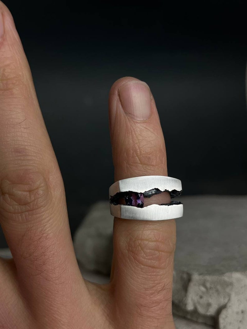 May include: A silver ring with a jagged, broken design. The ring has a purple gemstone set in the center of the break.