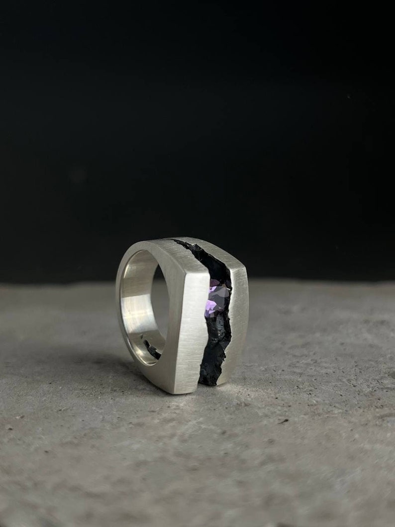 May include: A silver ring with a black crack down the center. A purple gemstone is set in the crack.