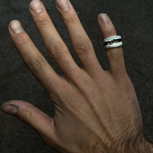 May include: A silver ring with a black band and a small purple stone set in the center. The ring is worn on the middle finger of a hand.