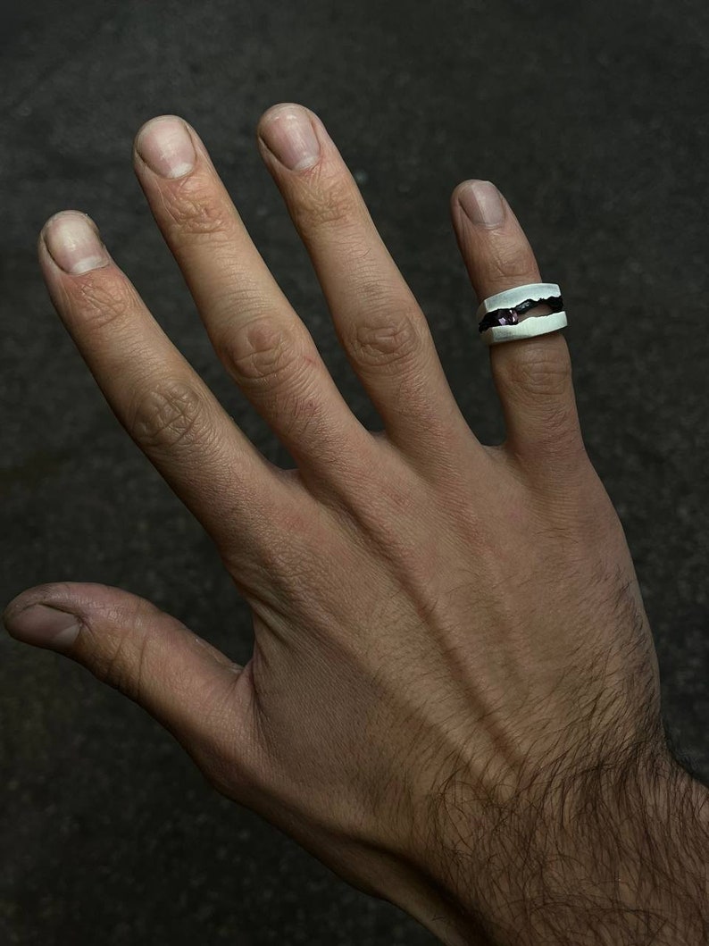 May include: A silver ring with a black band and a small purple stone set in the center. The ring is worn on the middle finger of a hand.