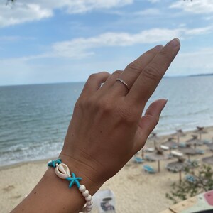 May include: A white beaded bracelet with turquoise starfish and a white seashell charm. The bracelet is worn on a person's wrist against a backdrop of a beach and ocean.