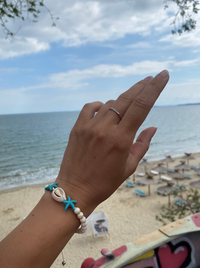May include: A white beaded bracelet with turquoise starfish and a white seashell charm. The bracelet is worn on a person's wrist against a backdrop of a beach and ocean.