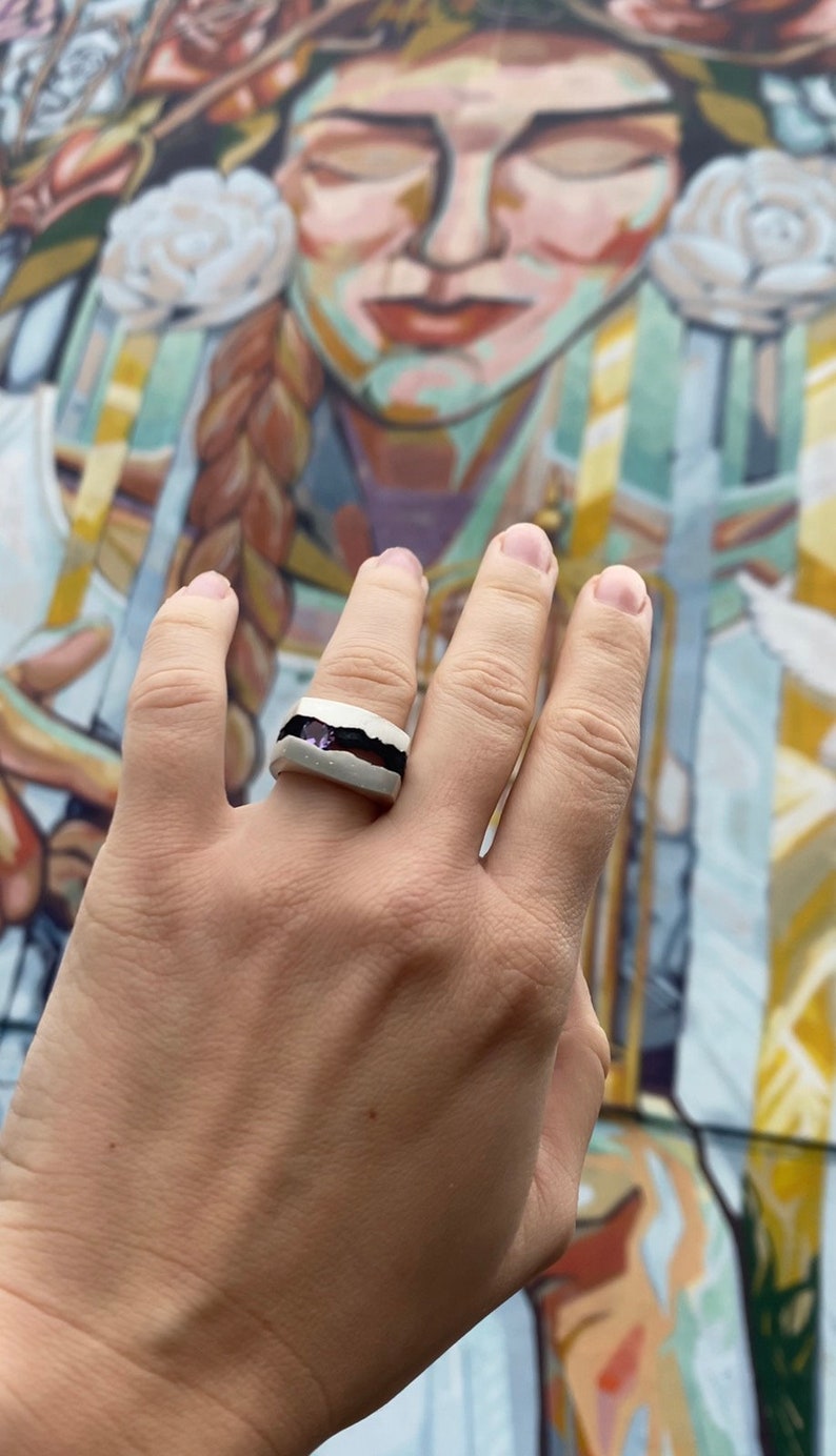 May include: A white ceramic ring with a black band and a purple stone inlay.