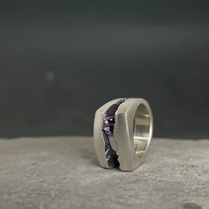 May include: A silver ring with a textured band and a purple gemstone set in the center.