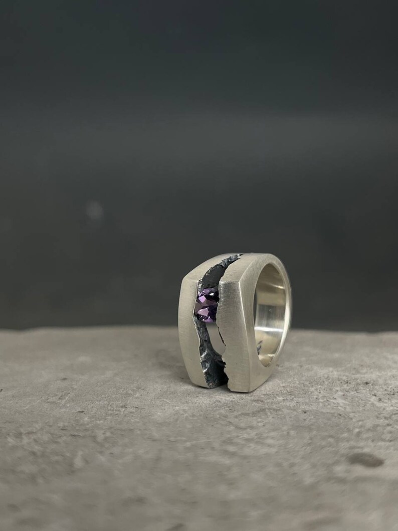 May include: A silver ring with a textured band and a purple gemstone set in the center.