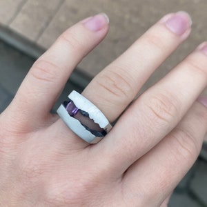 May include: A silver ring with a white band and a purple stone set in the center. The ring is designed to look like a broken piece of rock.