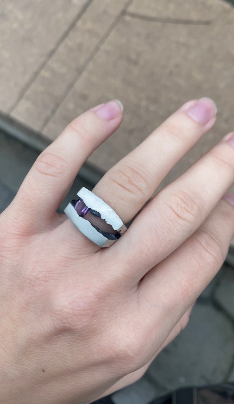 May include: A silver ring with a white band and a purple stone set in the center. The ring is designed to look like a broken piece of rock.