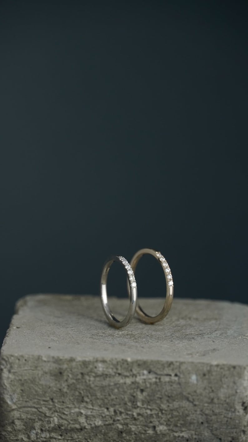 May include: Two thin wedding bands with small diamonds set in the band. The bands are on a gray concrete surface.