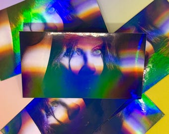 Op de afbeelding: Holografische dozen met een portret van een gezicht. De dozen hebben een reflecterende, iriserende afwerking met een regenboogeffect. Het gezicht is gedeeltelijk bedekt door het holografische ontwerp, waarbij de ogen het meest opvallende kenmerk zijn.