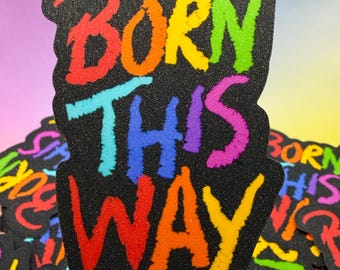 Op de afbeelding: Een zwarte sticker met de tekst "BORN THIS WAY" in regenboogkleuren. De tekst heeft een getextureerde, glitterachtige uitstraling. Andere stickers met hetzelfde ontwerp zijn zichtbaar op de achtergrond. Een paarse pincet houdt de sticker vast.