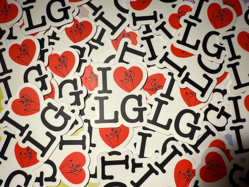 Op de afbeelding: Een verzameling witte stickers met de tekst "I LG" in zwarte letters, elk met een rood hart met een handillustratie. De stickers zijn verspreid, wat een visueel aantrekkelijk patroon cre&euml;ert.