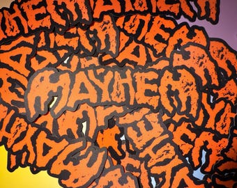 Op de afbeelding: Een verzameling oranje en zwarte patches met het woord "MAYHEM" in een gedurfde, distressed lettertype. De patches zijn verspreid over een kleurrijke achtergrond, wat een opvallend ontwerp creëert.
