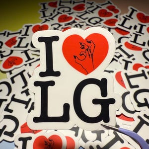 Op de afbeelding: Witte sticker met de tekst "I LG" in zwarte blokletters. De "I" wordt gevolgd door een rode hart met een zwarte handafbeelding. De stickers zijn verspreid over een gele ondergrond.