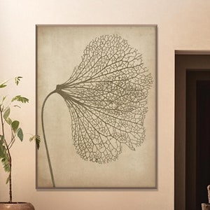 Op de afbeelding: Een beige muur met een grote ingelijste print van een bruin bladskelet. De print hangt boven een witte houten bank met een bruin stenen beeld erop.