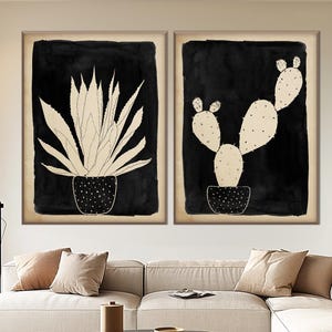 Könnte beinhalten: Zwei gerahmte schwarz-weiße botanische Kunstdrucke mit Kakteen und Agavenpflanzen. Das Kunstwerk hängt über einem beigefarbenen Ecksofa in einem modernen Wohnzimmer. Ein runder Couchtisch und ein Hocker befinden sich im Vordergrund.