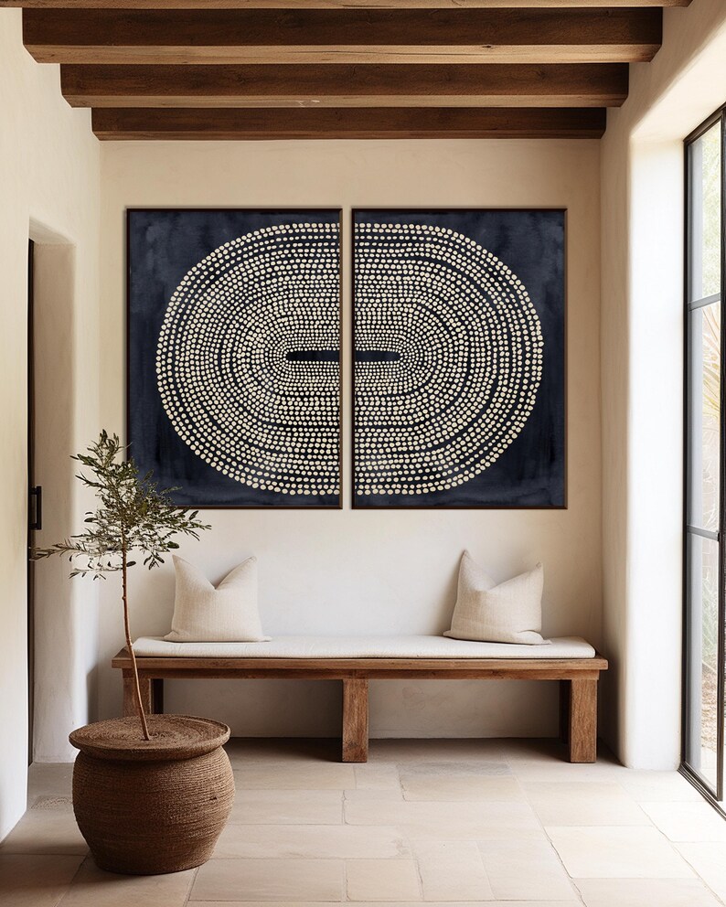 May include: Two abstract wall art pieces with a dark blue background and a white dotted oval design. The art hangs above a wooden bench with cream-colored pillows. A small tree in a woven pot sits nearby.