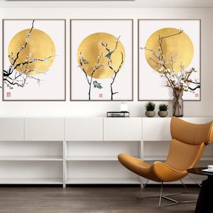 May include: Three framed art prints featuring a gold sun design with black branches, white flowers, and blue birds. The artwork is displayed above a white cabinet with a modern chair in a living room setting.
