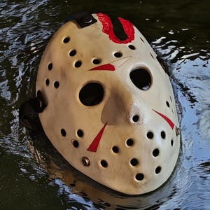 Könnte beinhalten: Eine cremefarbene Hockeymaske mit schwarzen Augenlöchern und zahlreichen kleinen, gleichmäßig verteilten Löchern. Rote Markierungen sind auf der Maske vorhanden, die teilweise in Wasser getaucht ist. Die Maske hat schwarze Riemen.