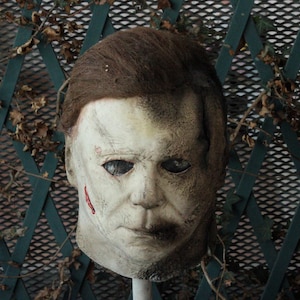 Può includere: Una maschera in lattice a testa intera che raffigura un volto umano pallido e segnato dal tempo, con capelli castano scuro, orbite profonde e un piccolo segno rosso a forma di taglio sulla guancia destra. La maschera ha una texture invecchiata.