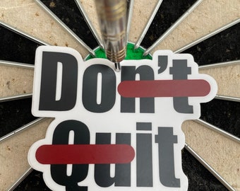 Op de afbeelding: Een witte sticker met de woorden "Don't Quit" in zwarte tekst. Het woord "Quit" is doorgestreept met een rode lijn. De sticker zit op een dartbord.
