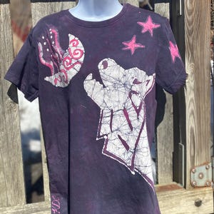 May include: Dark purple t-shirt with a batik design featuring a bear, stars, and abstract shapes in white and pink. The shirt is displayed on a mannequin. The artist's initials, KDC, are visible on the lower left side.