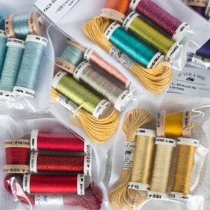 Könnte beinhalten: Nahaufnahme von mehreren Spulen mit Stickgarn in verschiedenen Farben, darunter Rot, Blau, Gelb und Grün. Die Spulen sind mit dem Markennamen "Au Ver a Soie" und dem Farbnamen beschriftet. Die Spulen sind in durchsichtigen Plastiktüten verpackt.