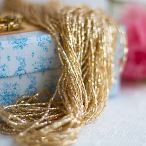 Könnte beinhalten: Nahaufnahme einer großen Spule mit goldfarbenen Perlen. Die Perlen sind in einem lockeren, fließenden Haufen angeordnet, und die Spule liegt auf einer blau-weiß gemusterten Schachtel.