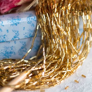 Può includere: Primo piano di un mucchio di perline di vetro dorate. Le perline sono piccole e cilindriche e sono disposte in un mucchio sciolto e fluido. Le perline sono sparse su una superficie bianca.