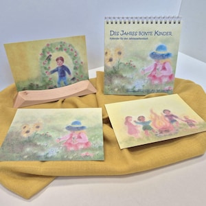 Könnte beinhalten: Eine Sammlung von Kunstdrucken für Kinder und ein Spiralbuchkalender. Der Kalender und die Drucke zeigen Aquarellillustrationen von Kindern in der Natur. Der Text auf dem Kalender lautet "Des Jahres Bunte Kinder". Die Artikel werden auf einem gelben Tuch präsentiert.