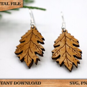 May include: A pair of wooden earrings shaped like oak leaves. The earrings are a light brown color and have a detailed design. The earrings are hanging from silver hooks.